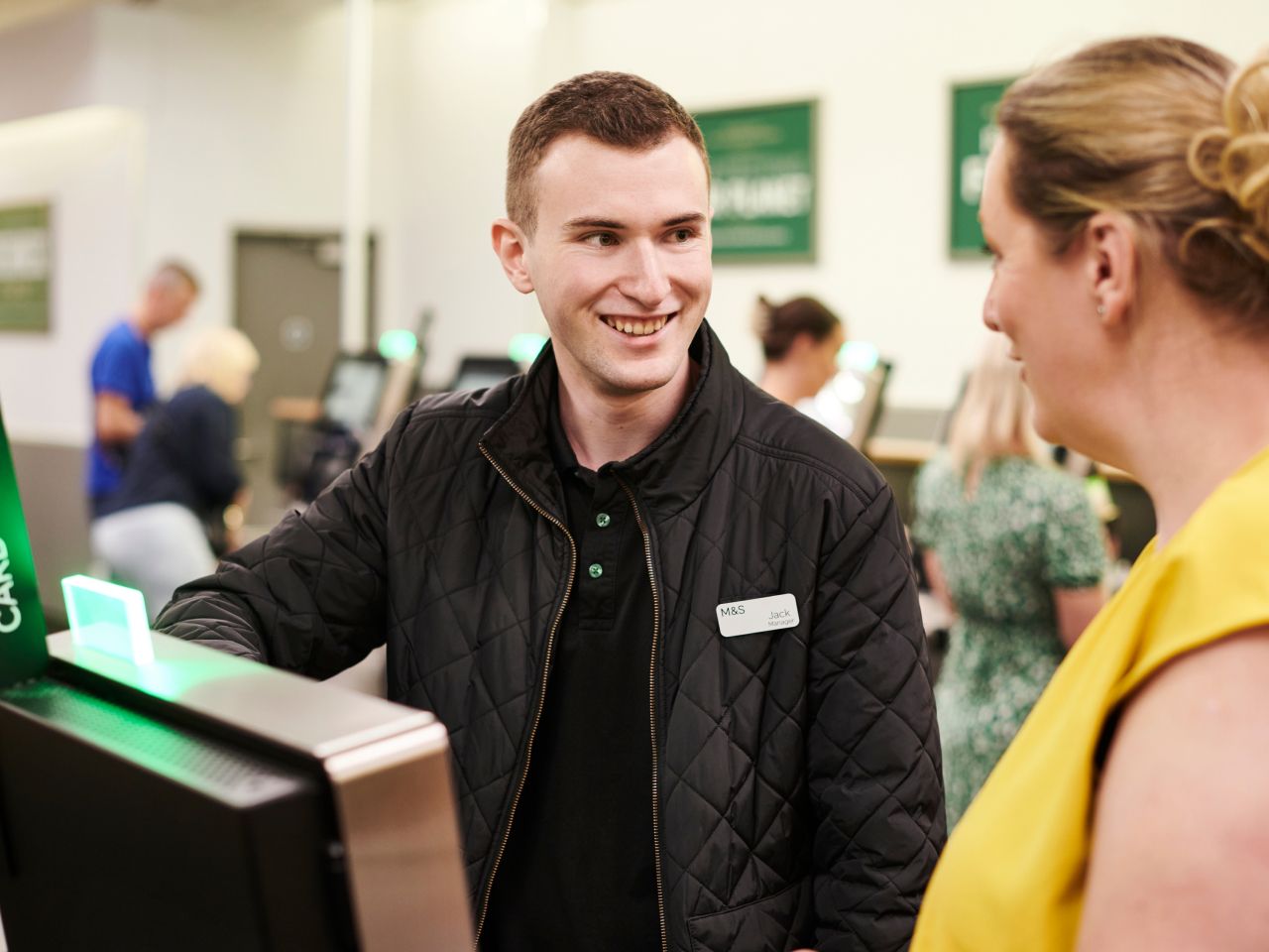 A Marks & Spencer employee wearing a quilted jacket, smiling and assisting a customer at the checkout area.