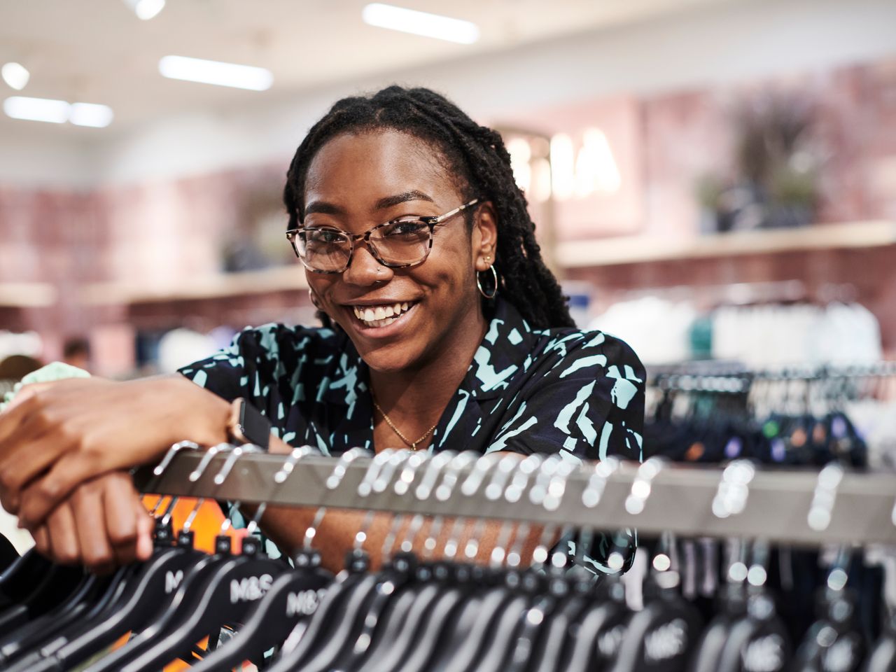 A smiling retail employee with glasses and dreadlocks leaning on a clothes rack in a Marks & Spencer store, wearing a patterned shirt.