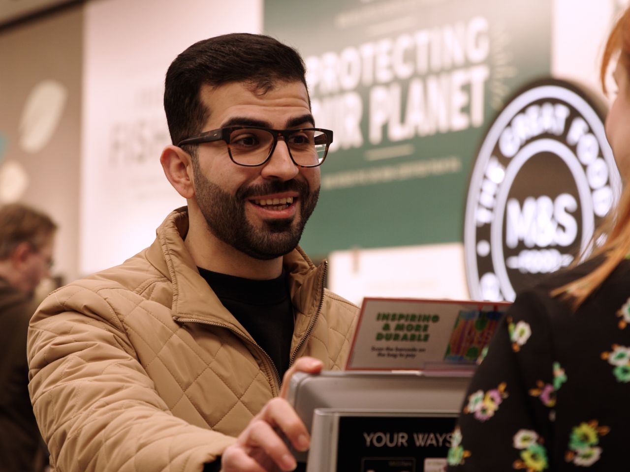 A man in glasses and a quilted jacket smiling while interacting with a Marks & Spencer employee at a checkout counter.