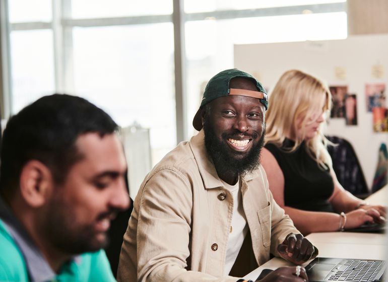 A cheerful man wearing a cap and jacket smiling while working alongside colleagues in a bright office environment.
