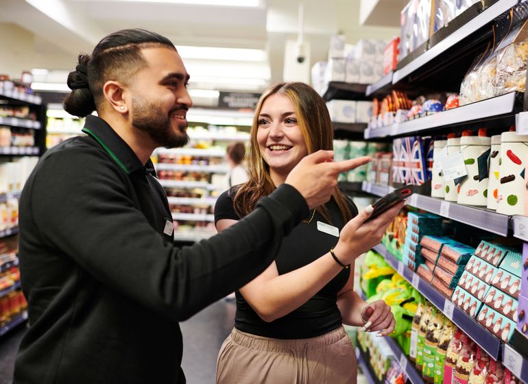 Two Marks & Spencer employees smiling as they discuss products on a store shelf, with one pointing and the other holding a tablet.
