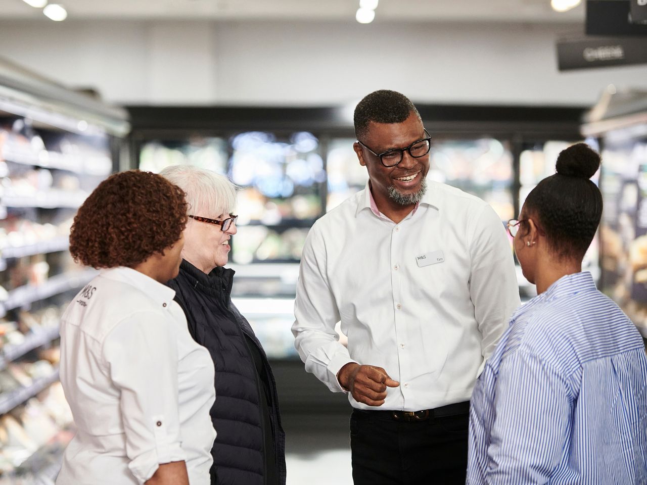 A group of Marks & Spencer employees standing in an aisle, smiling and engaged in conversation in a store setting.