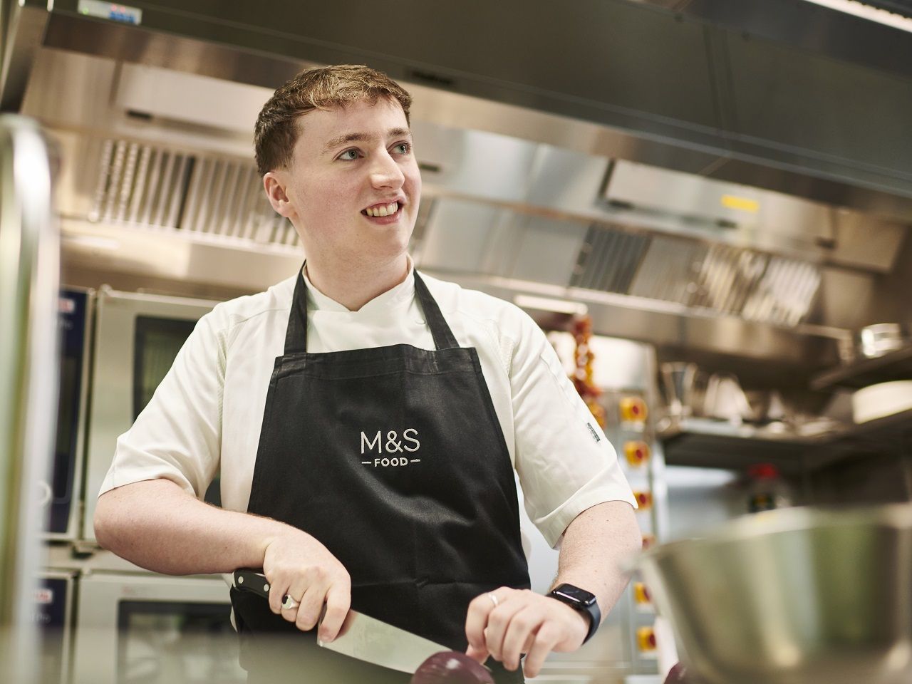 A Marks & Spencer chef in a professional kitchen, smiling while chopping vegetables, wearing a white chef's coat and a black M&S Food apron.