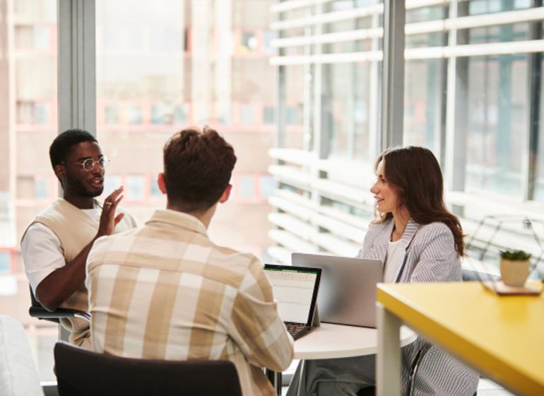 A diverse group of three colleagues sits around a table in a modern office setting. One person gestures while speaking, and the others listen attentively with open laptops in front of them. 