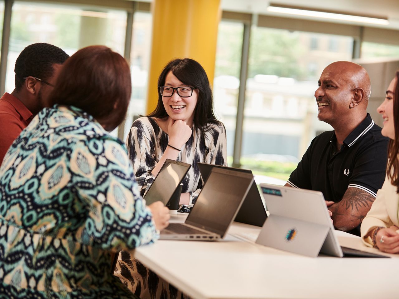 A group of colleagues sitting around a table with laptops, smiling and engaged in conversation in a modern office setting.