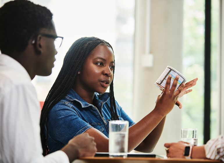 A Marks & Spencer employee holding a packaged product, engaged in a discussion with colleagues around a table in a meeting area.