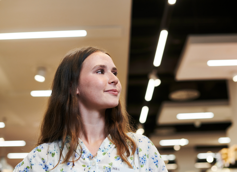 A Marks & Spencer employee in a floral blouse, standing confidently and looking to the side in a well-lit store environment.