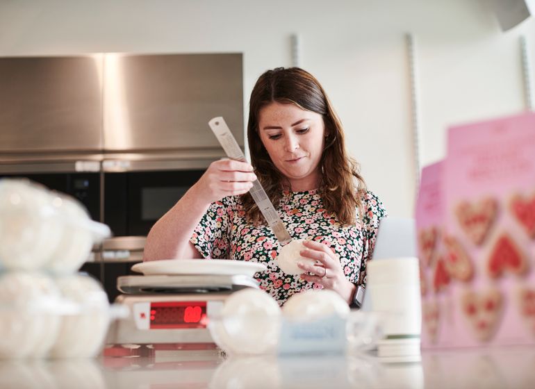 A woman in a floral shirt carefully measuring ingredients in a kitchen at Marks & Spencer, surrounded by baking supplies.