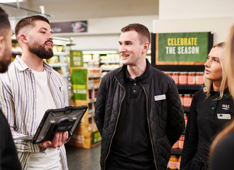 A group of Marks & Spencer employees standing in a store aisle, engaged in discussion, with one holding a tablet. 