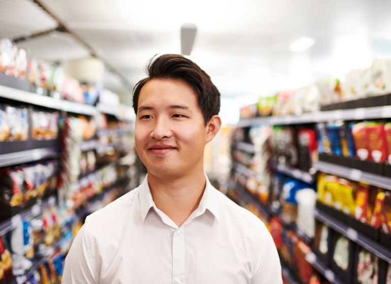 A Marks & Spencer employee standing in an aisle, smiling and looking to the side, surrounded by shelves stocked with products.