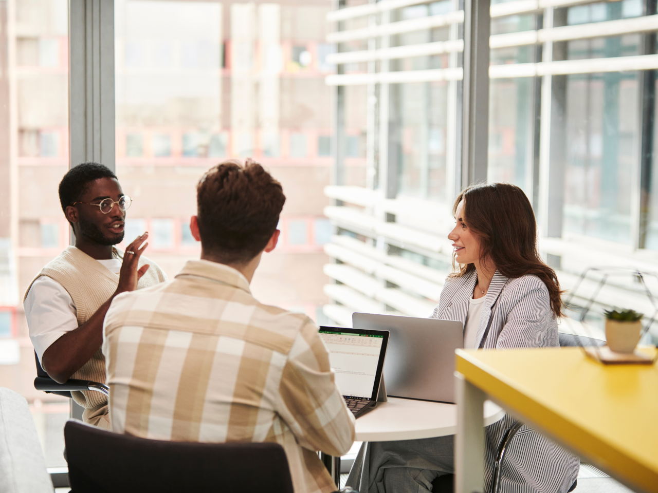 A diverse group of three colleagues sits around a table in a modern office setting. One person gestures while speaking, and the others listen attentively with open laptops in front of them.