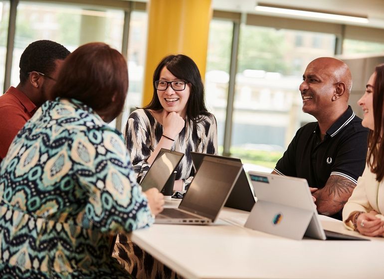 A group of colleagues gathered around a table, smiling and discussing work with laptops open in a modern office setting.