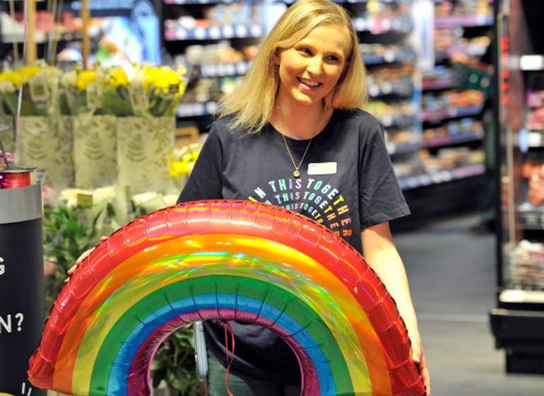 A smiling Marks & Spencer employee holding a rainbow-shaped balloon in a store aisle, with flowers and products displayed in the background.