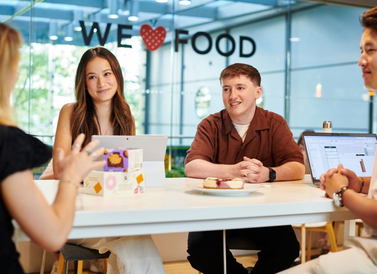 A group of four colleagues sits around a table in a bright, modern office. They appear engaged in a casual meeting, with two of them looking towards the person speaking. A laptop and some food items, including packaged snacks, are on the table.