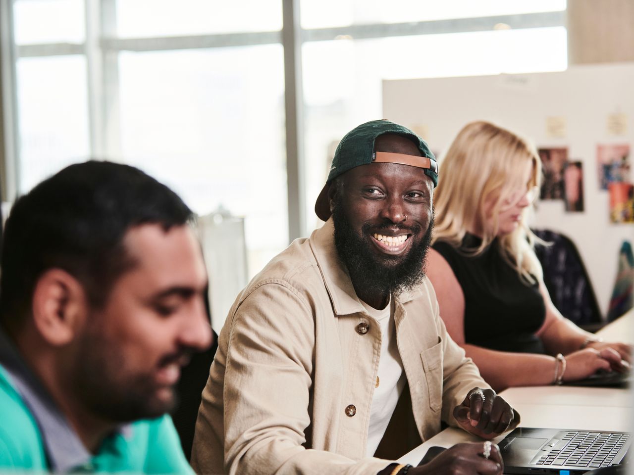 A cheerful man wearing a cap and jacket smiling while working alongside colleagues in a bright office environment.