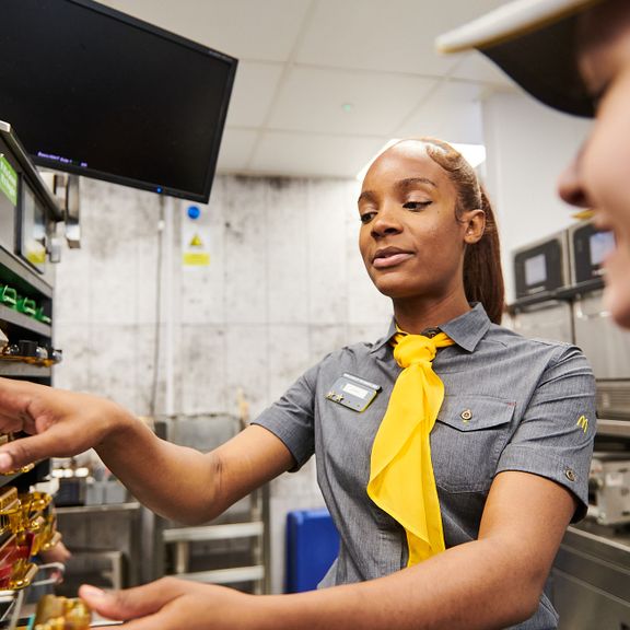 Two employees in a fast-food kitchen, one is handling a food station.