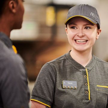 Two individuals in grey uniform in a fast-food kitchen, one is smiling at the other.