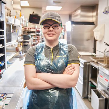 A person standing in a fast-food kitchen in a grey uniform and a cap with their arms crossed.