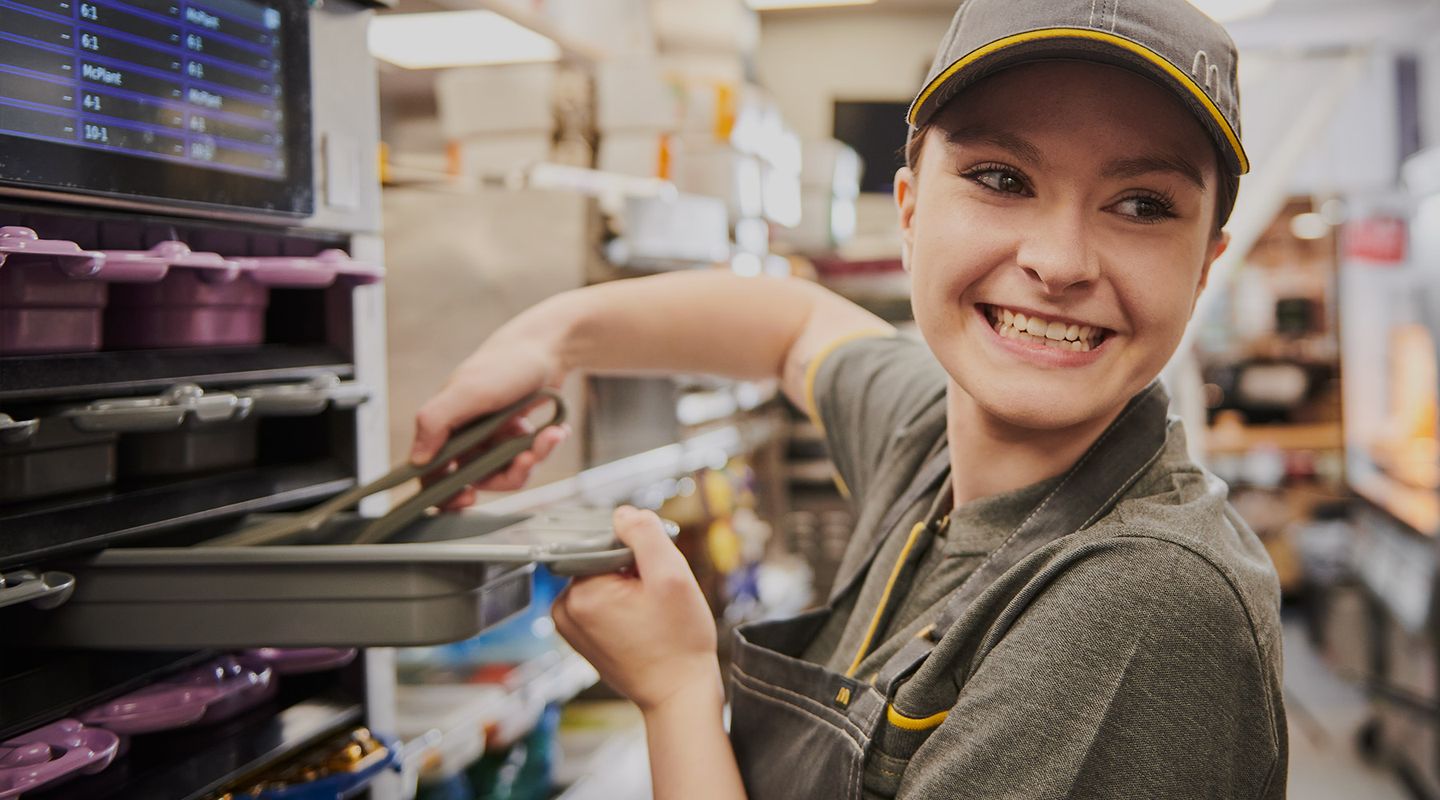 A smiling fast-food worker in uniform uses tongs in a busy kitchen, standing in front of shelves filled with supplies.