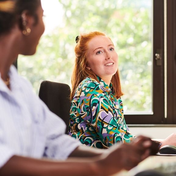 Two people in an office setting, sitting side by side at a desk, both are in a cheerful mood.