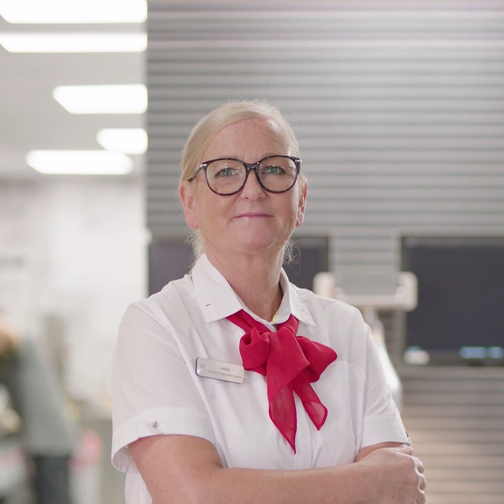 Employee in white uniform with red scarf, arms crossed, in a fast-food restaurant.