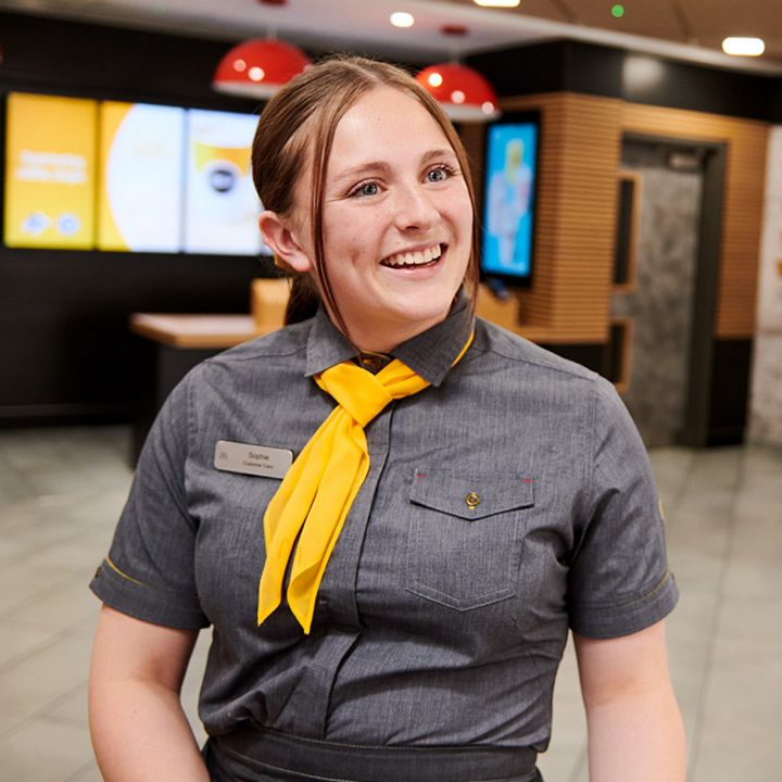 A cheerful woman standing in a fast-food restaurant in a grey uniform with a yellow necktie.