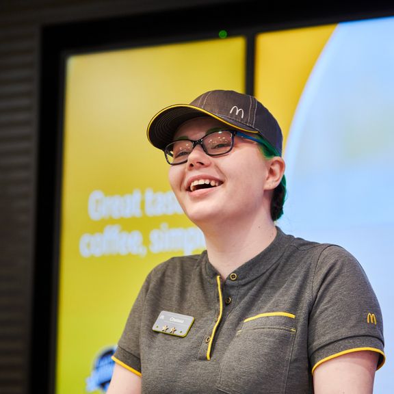 A person wearing a grey uniform and a cap, standing in front of a yellow digital menu board.