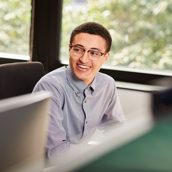 A person with glasses smiling, sitting at a desk in an office setting.