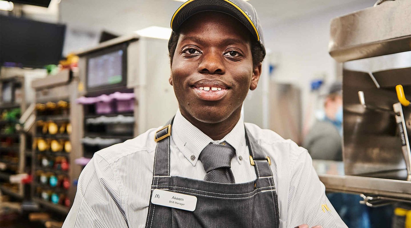 An individual wearing a grey uniform and a cap, smiling, with their arms crossed, standing in a fast-food kitchen.