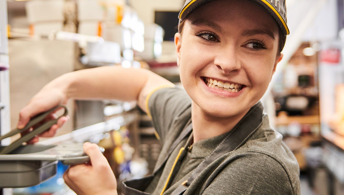 A smiling fast-food worker in uniform uses tongs in a busy kitchen, standing in front of shelves filled with supplies.