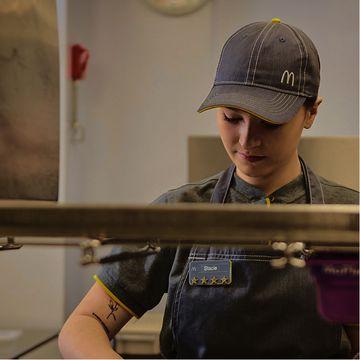 An employee wearing a grey uniform preparing food in a fast-food kitchen, with a hat on.