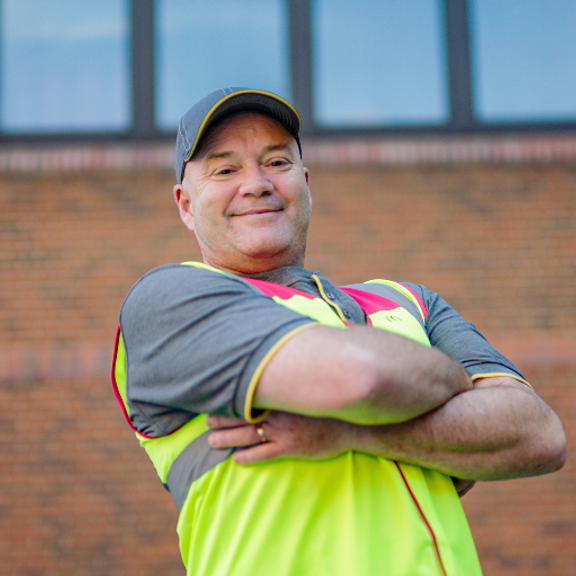 An employee smiling with a high-visibility jacket, with their arms crossed, standing outside.