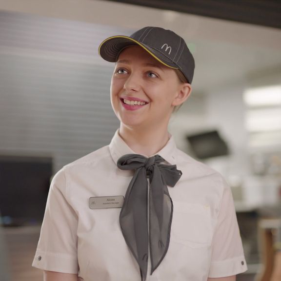 A smiling woman in a McDonald's uniform with a gray necktie and cap stands in a well-lit restaurant setting.
