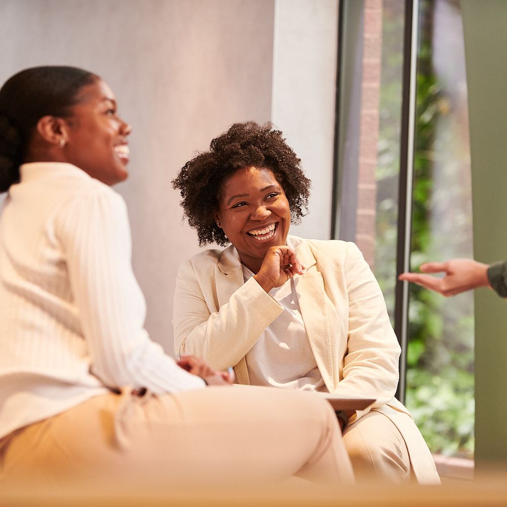 Two individuals smiling and laughing, sitting in an office setting, they are in conversation with a third person.