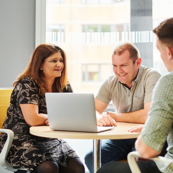 Three individuals collaborating around a table in a bright office space, one using a laptop.