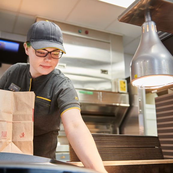 Worker in uniform at a fast-food counter with a paper bag, under a heat lamp.