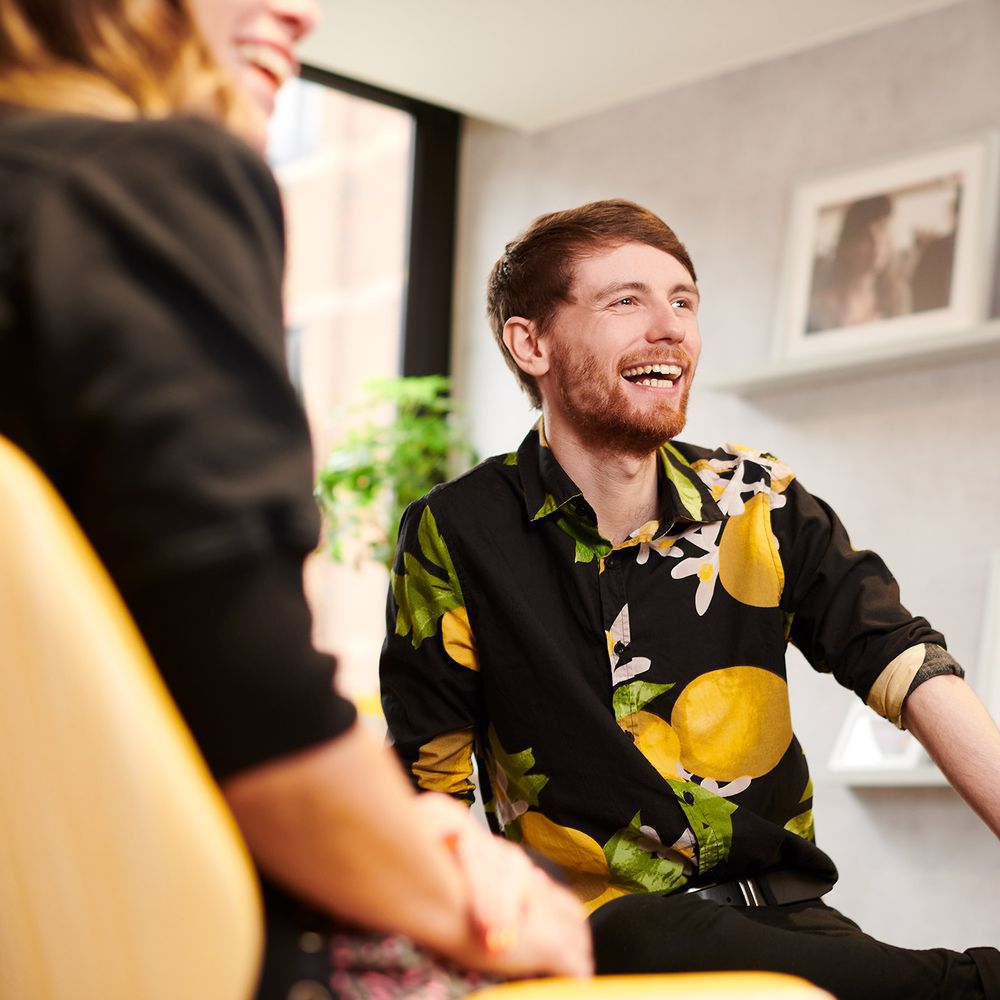 Two cheerful individuals, smiling in an office setting. Both are seated.