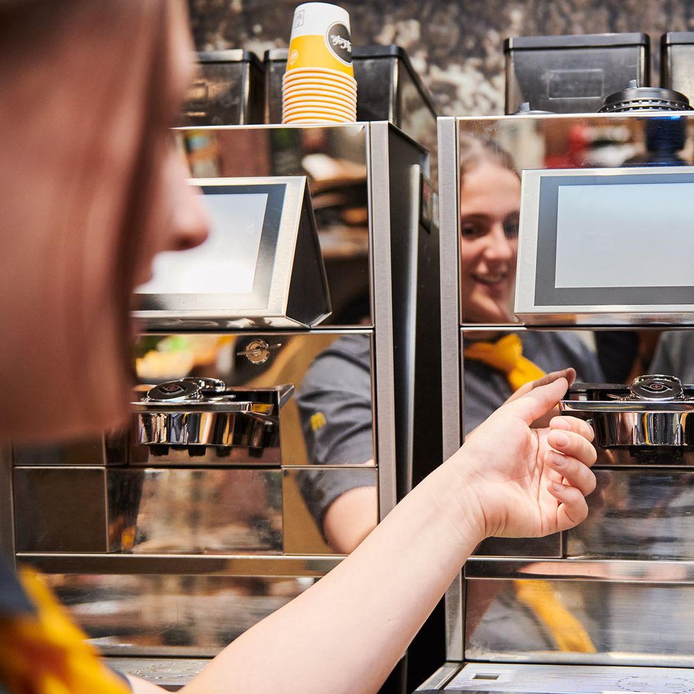 A woman using a coffee machine with a touchscreen interface, with disposable cups stacked on top. The person's reflection can be seen in the machine.