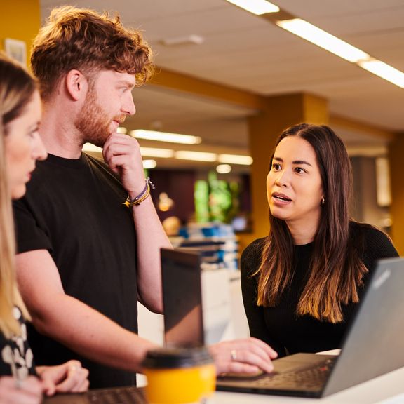 Three individuals gathered around a laptop collaborating and standing in a bright office setting.