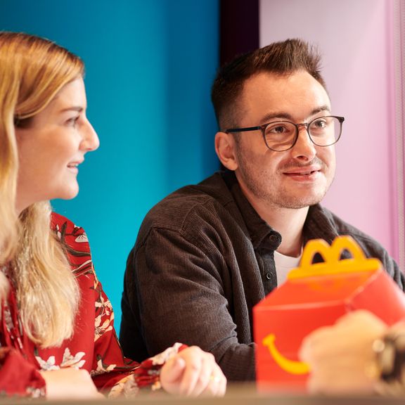 Two individuals at a table, both smiling, in an office setting. There is food packaging on the table.