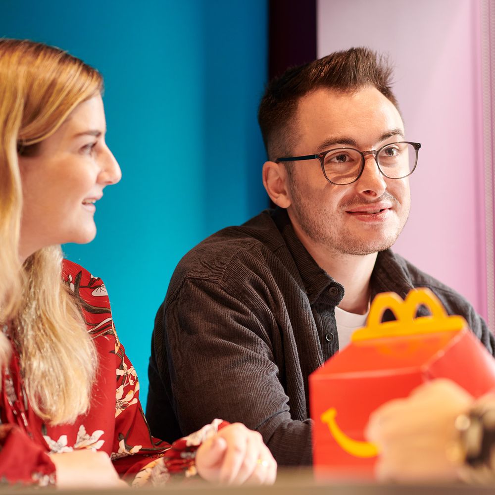 Two individuals at a table, both smiling, in an office setting. There is food packaging on the table.