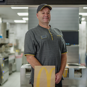 A man smiling in grey uniform, standing in a fast-food kitchen with his arms folded down, with food packaging in front of him.