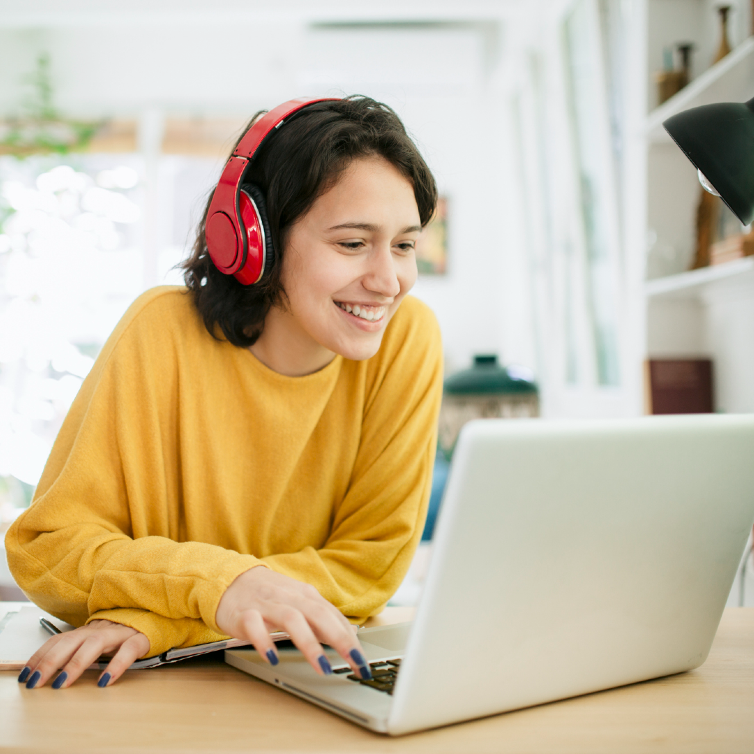 A person in a yellow shirt, smiling, using a laptop with red headphones on.