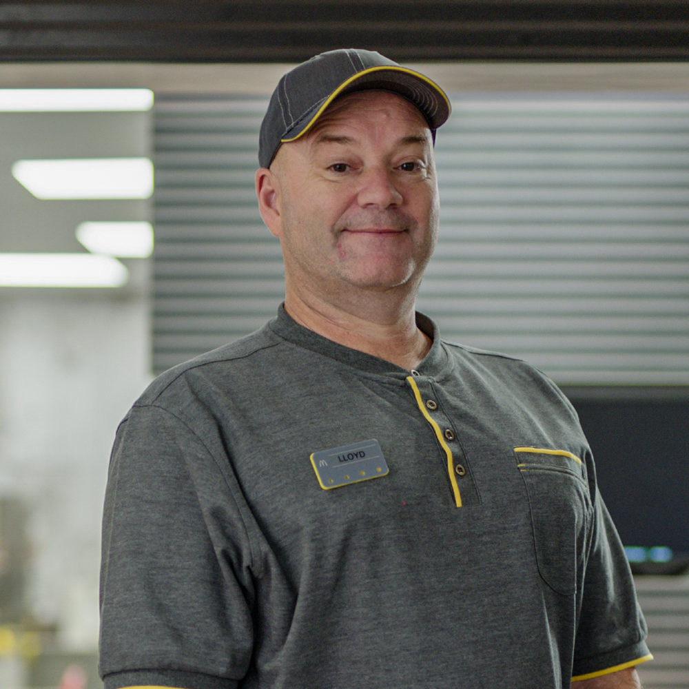 A man smiling in grey uniform, standing in a fast-food kitchen with his arms folded down.