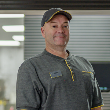 A man smiling in grey uniform, standing in a fast-food kitchen with his arms folded down.