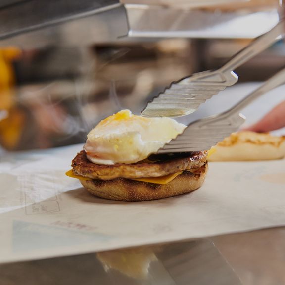 Tongs place a cooked egg on a sausage patty and cheese, all on an English muffin, in a busy fast-food kitchen.