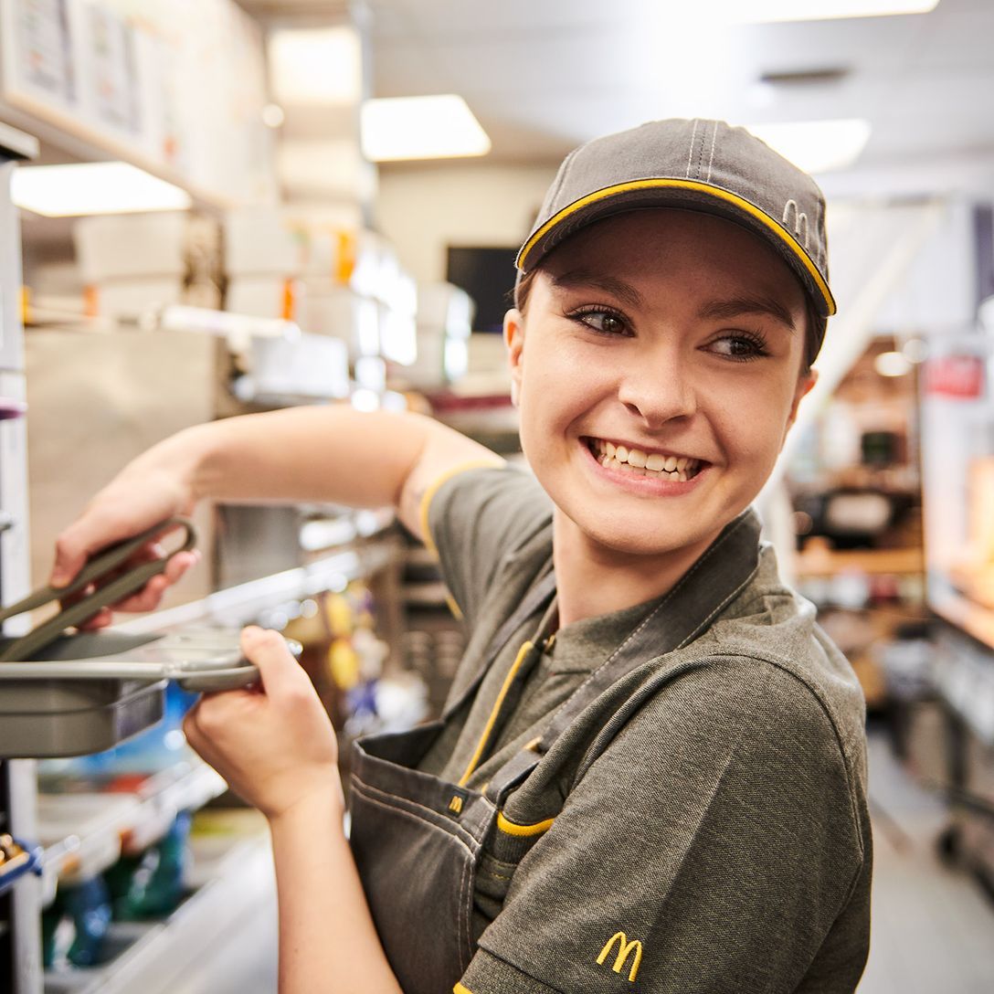 A smiling fast-food worker in uniform uses tongs in a busy kitchen, standing in front of shelves filled with supplies.