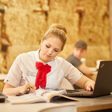 A woman in a white shirt and red tie sitting at a table with a laptop, writing on a page.