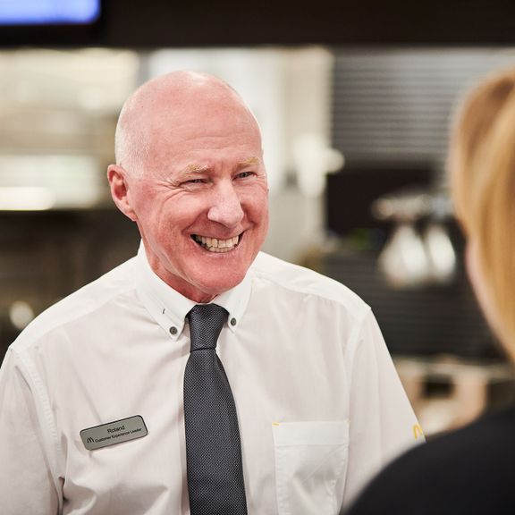Employee wearing a white shirt, black tie, smiling at someone out of the frame.