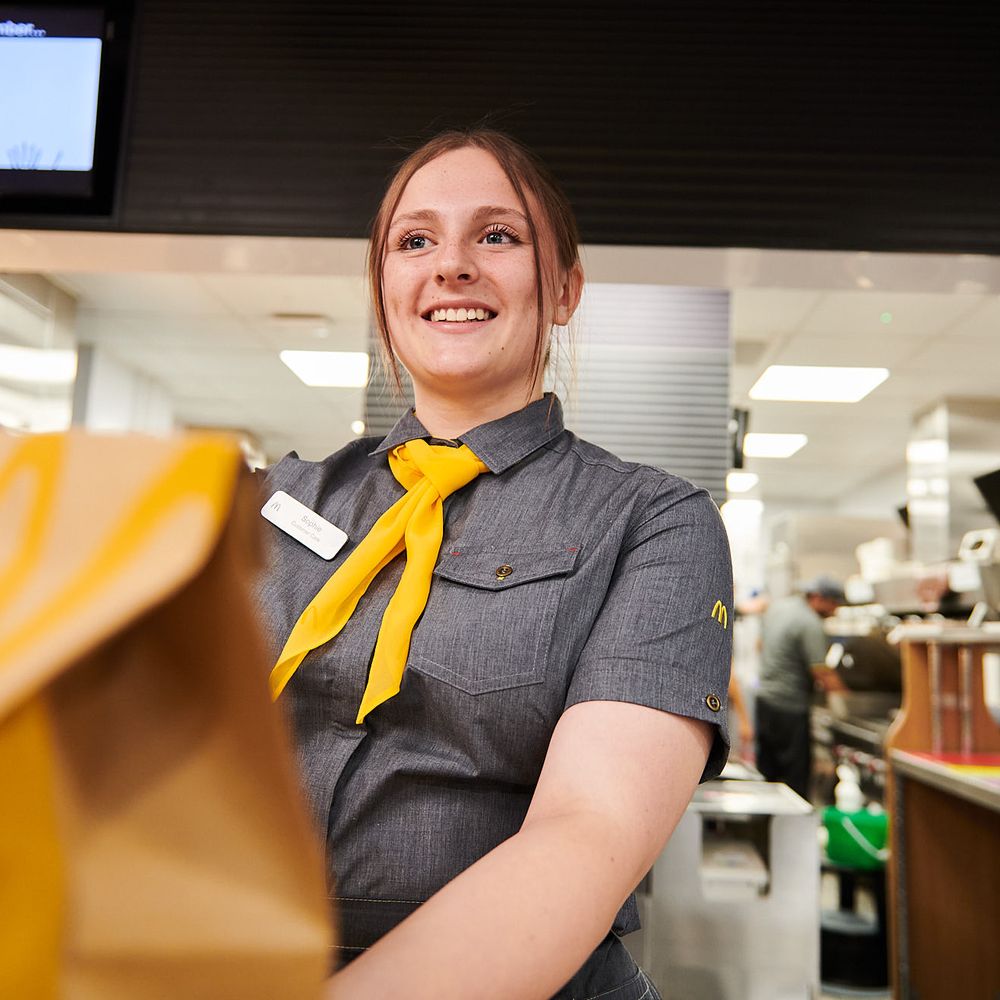 A cheerful woman in a uniform with a yellow necktie hands over a paper bag, at a fast-food restaurant counter.